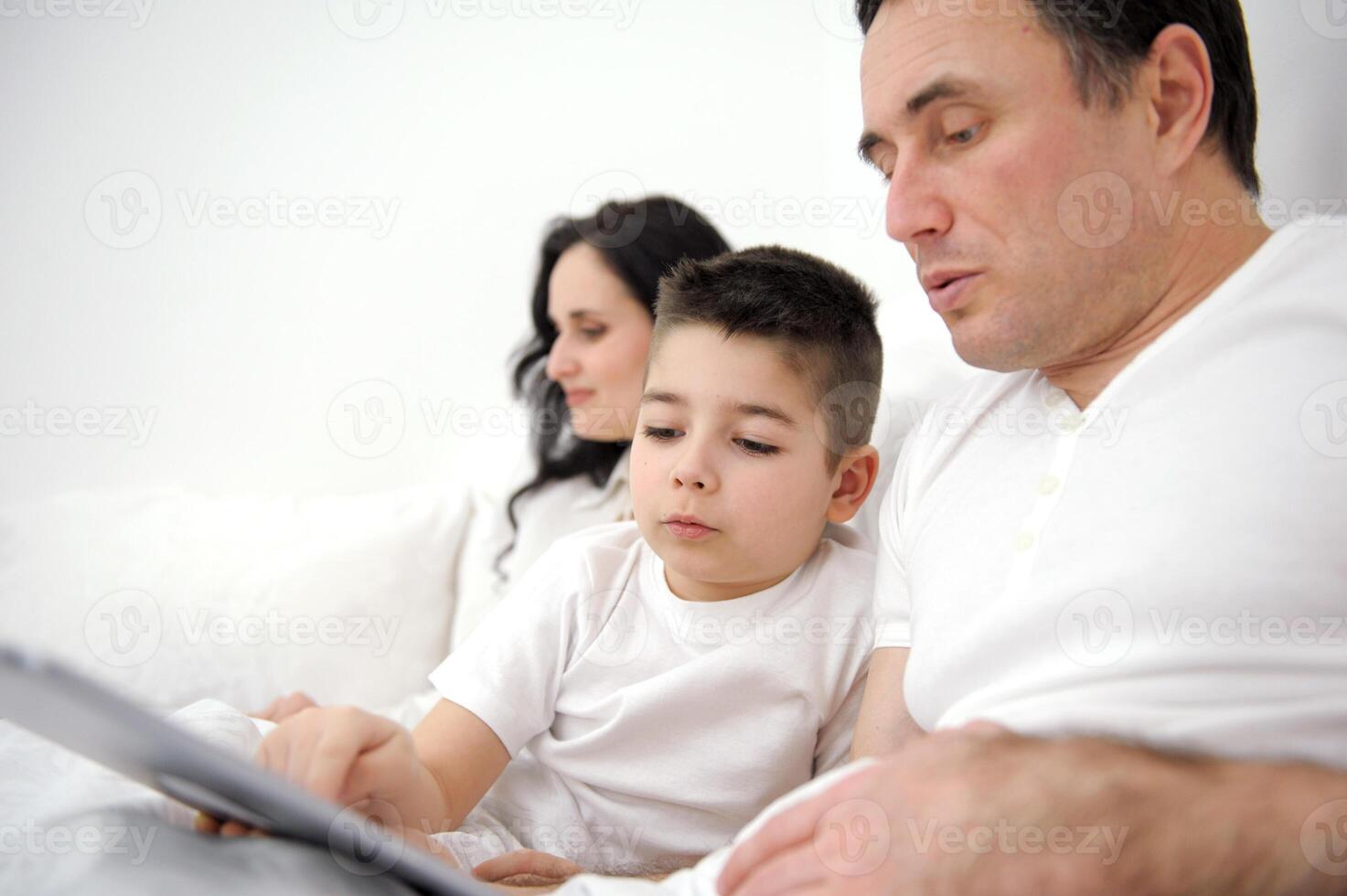 family, fatherhood, technology and people concept happy father and little son with tablet pc computer sitting in bed at home mom woman working in the background online on computer laptop photo