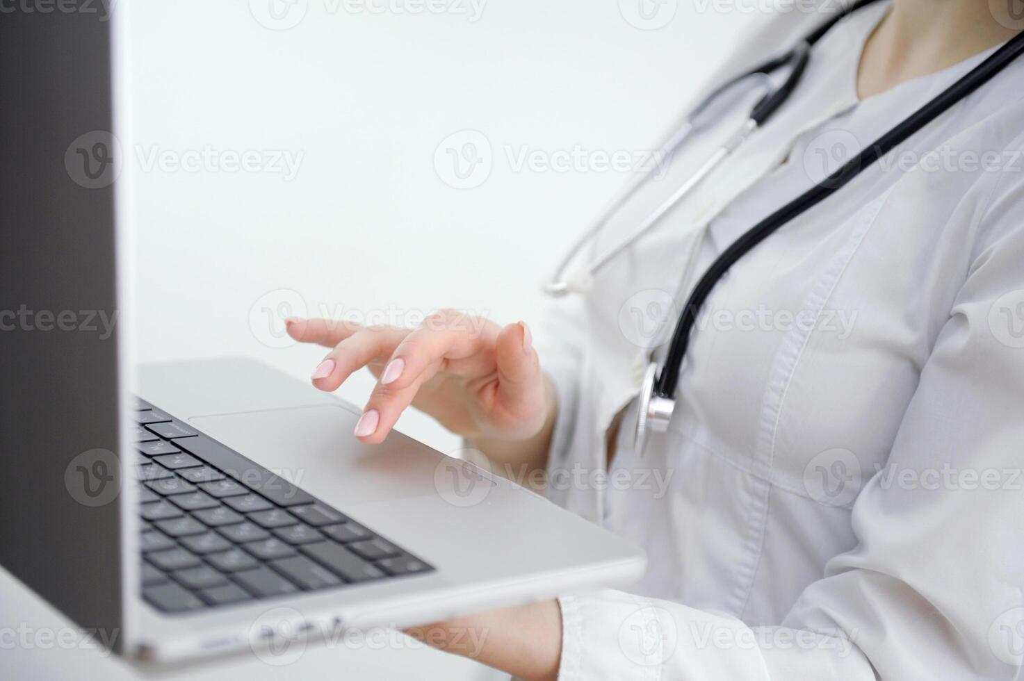 Close up of unknown female doctor sitting at the table near the window in hospital and typing at laptop computer photo