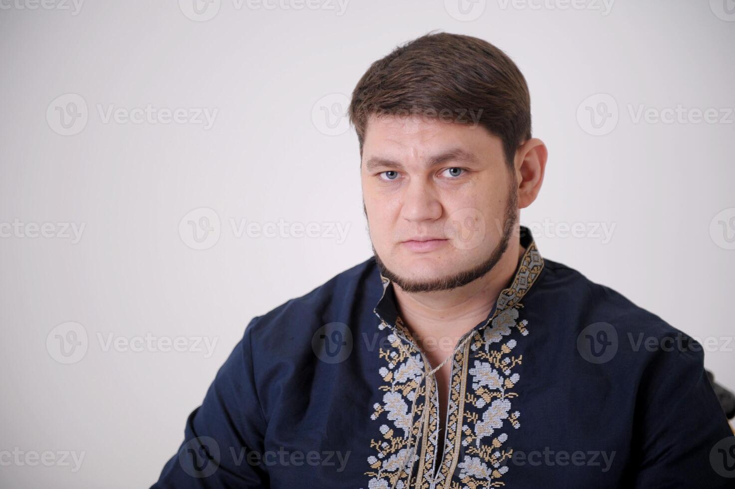 stern look of blue eyes handsome man in Ukrainian embroidered shirt looks straight into frame he has light beard compressed lips thick hair a blue shirt and embroidery on a white background close-up photo