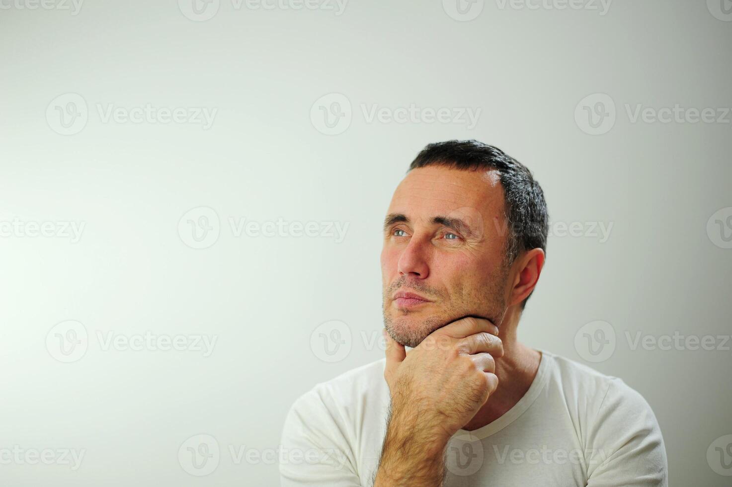 Strict unshaven man looks into the frame Hold your chin with your hand and think He has a strong piercing blue eyes he is a studio on a white background Problems of experiencing a decision photo