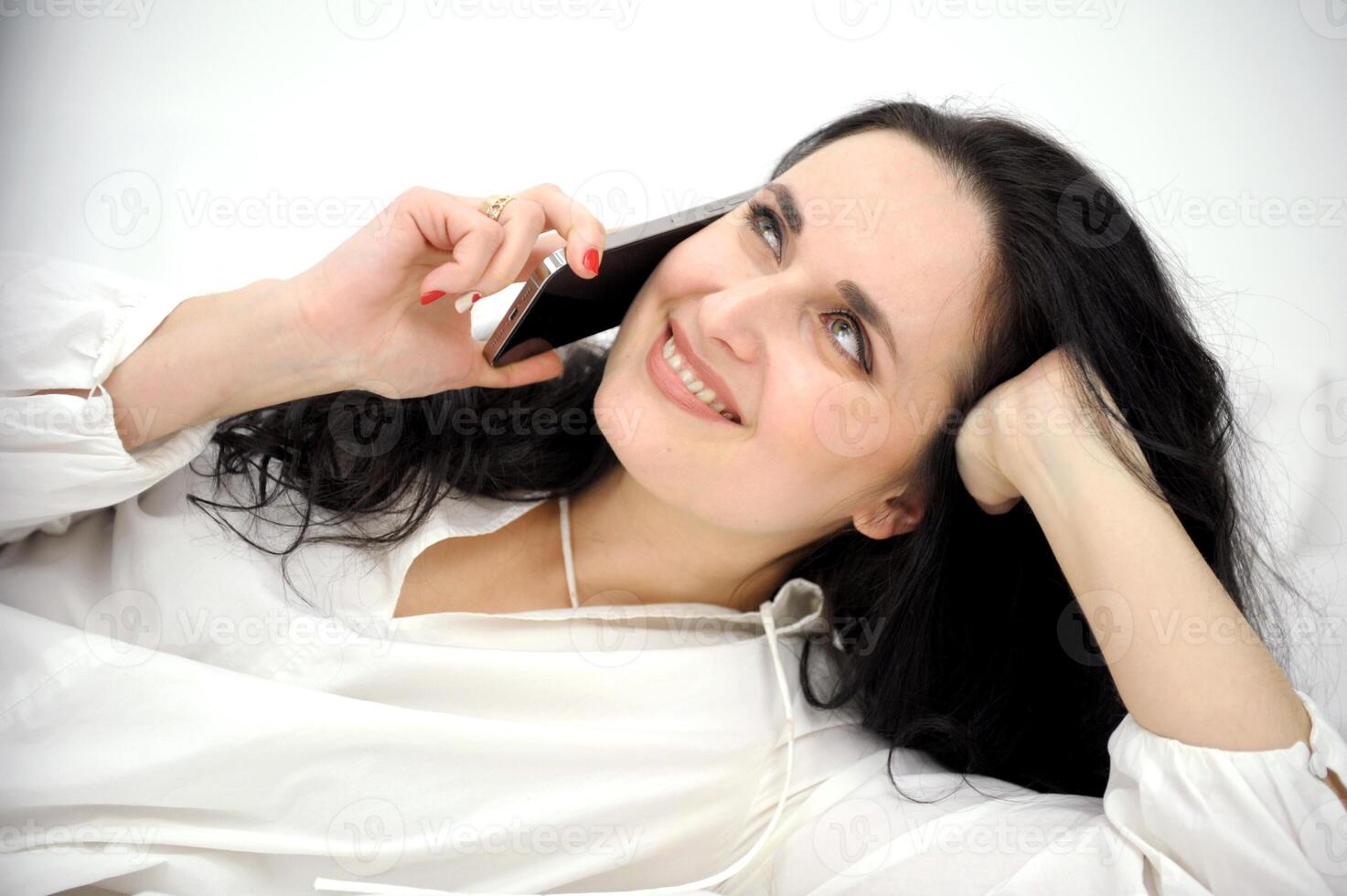brunette with mobile phone Close-up of happy sensual asian girl lying in bed and using mobile phone. Woman smiling cheerful as looking at mobile phone, resting and texting message on social network photo