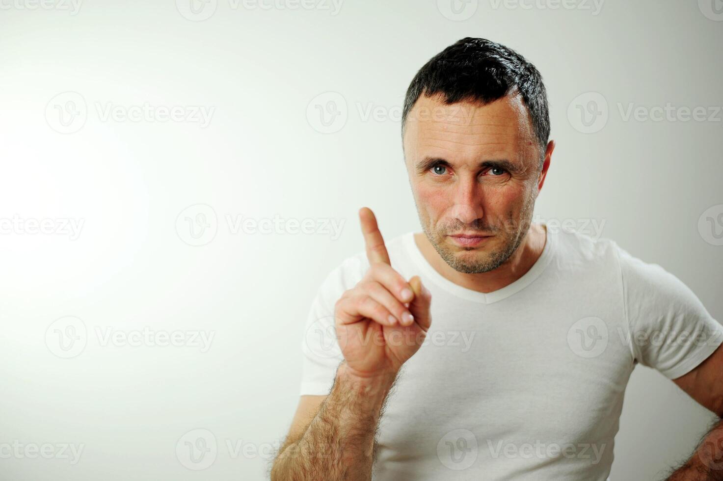 Serious man looking into frame raised his finger up Adult unshaven handsome man Strictly stares into the frame billboard banner Open wide blue eyes white background studio Advertising space for text photo