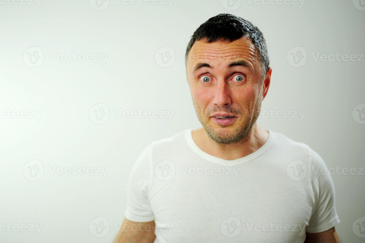 attractive unshaven man astonished amazed in shock surprise face expression and shock emotion Blue eyes black hair with gray White t-shirt in the studio photo