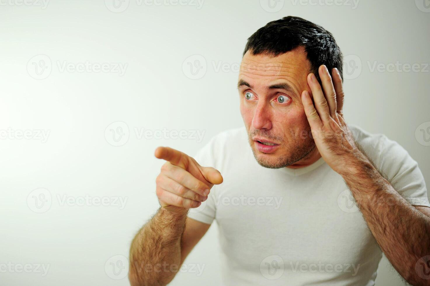 Adult unshaven handsome man shows his finger With one hand holding his head, as if looking billboard banner Open wide blue eyes white background studio Advertising space for text photo