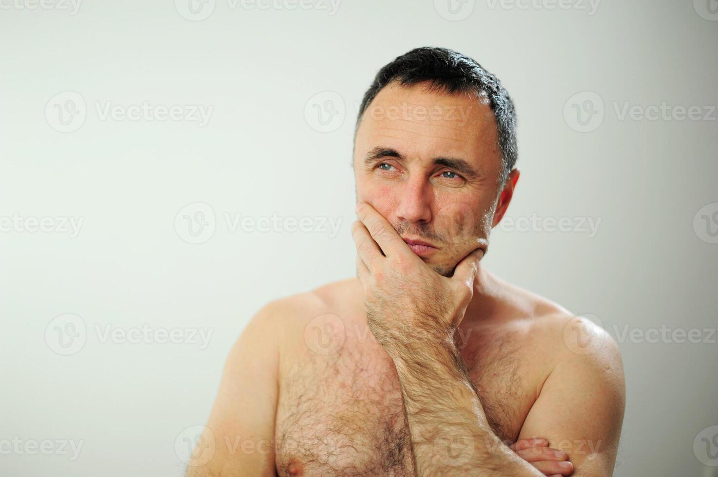Unshaven man touches his beard with his hand and looks away as if he is about to shave Cream cologne man with a hairy chest and hairy arms on a white background with black hair with gray hair photo