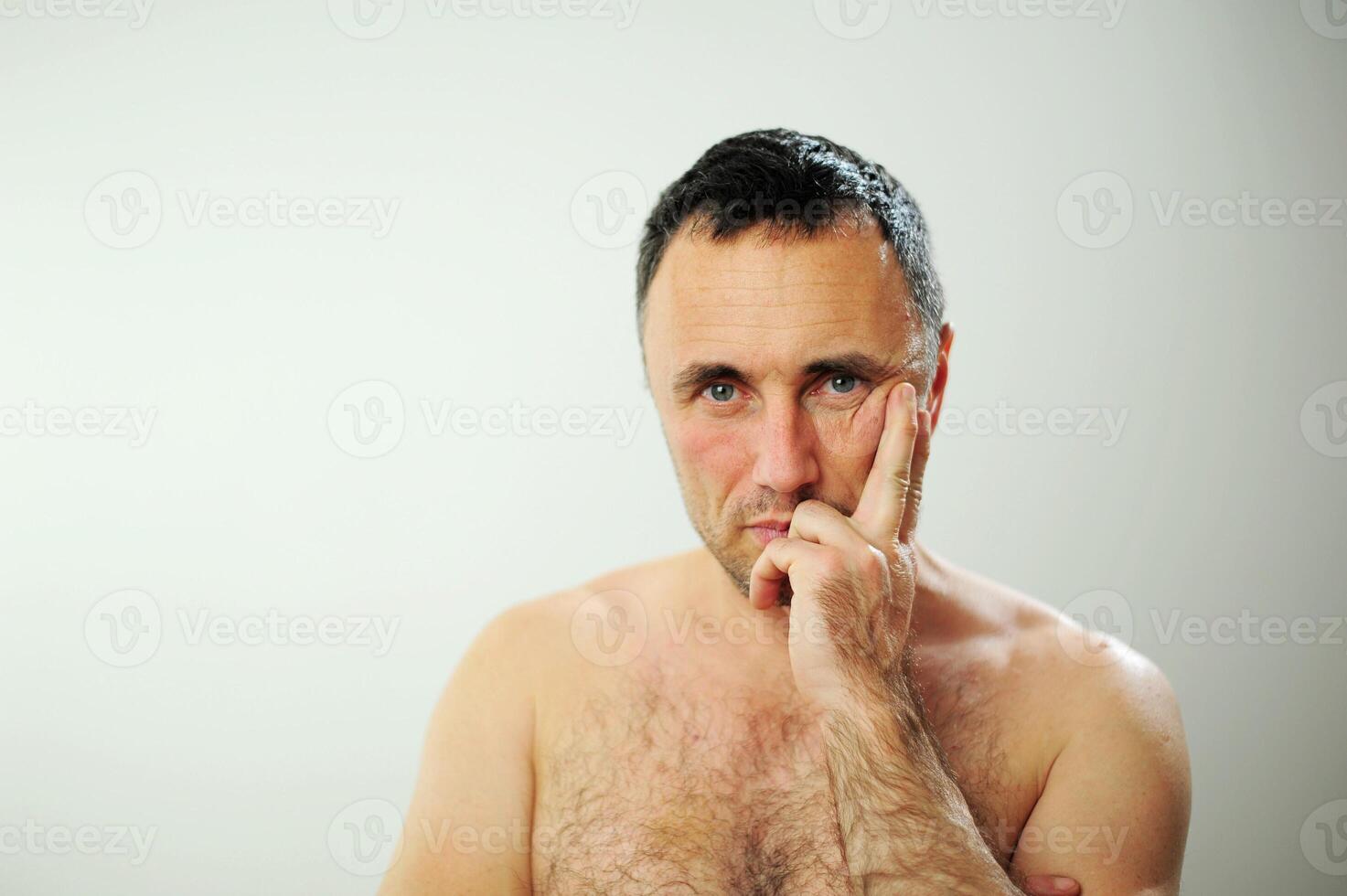 male adult with a naked torso looks into the frame He doubts or his tooth hurts he pressed his finger to his cheek dark hair on a white background there is a place for advertising foreground photo