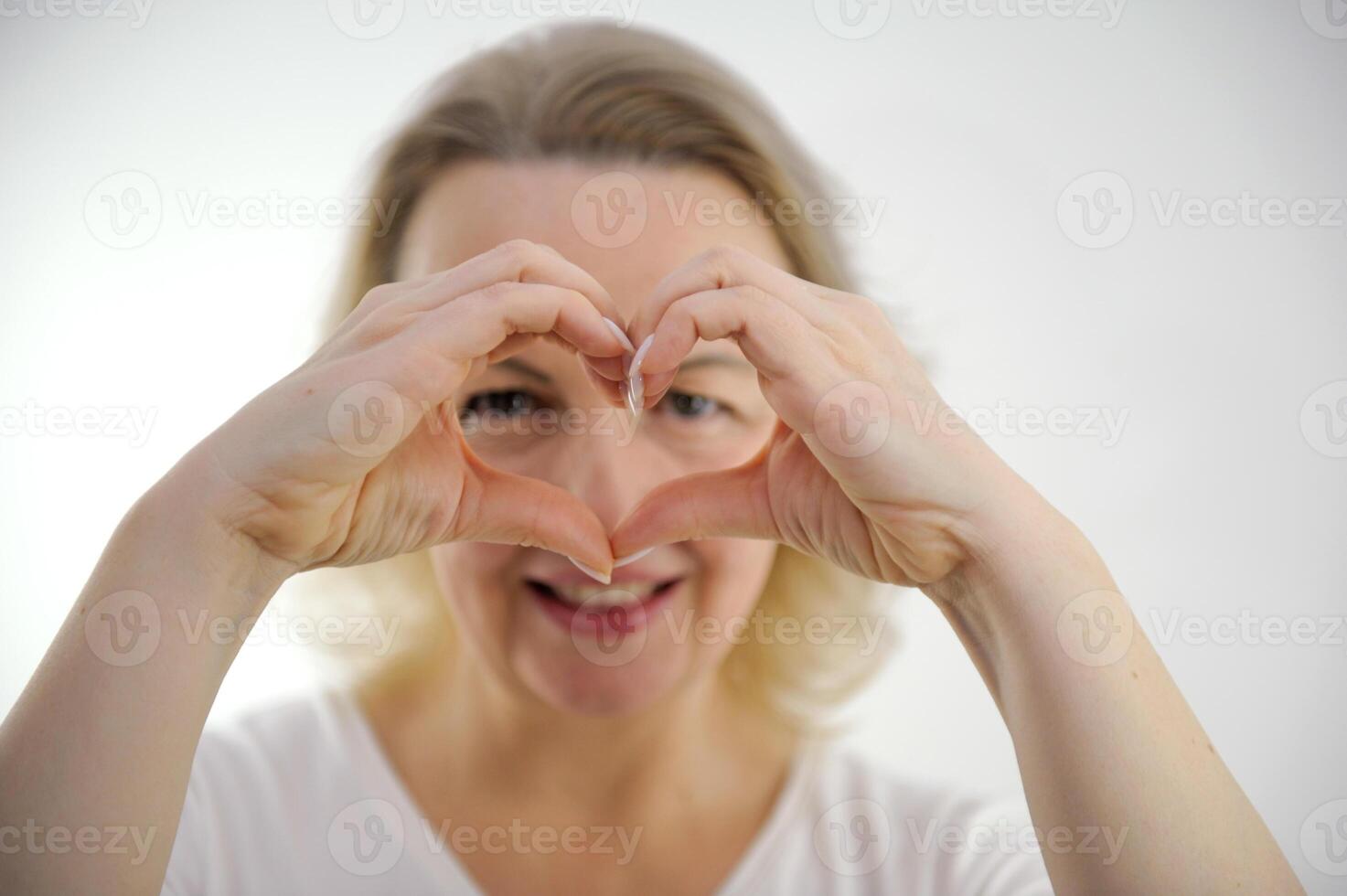 middle-aged woman showing heart with hands looks through hole smiles love expectations recognition tenderness beauty hands in sharpness selective sharpness on white background female feelings photo