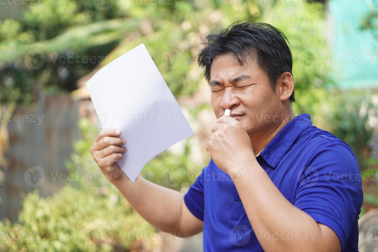 Asian man feel hot, unwell, use menthol inhaler to smell, hold paper to fan for relief symptom from hot weather. Concept, self first aid to protect from heat stroke. Refreshing. photo
