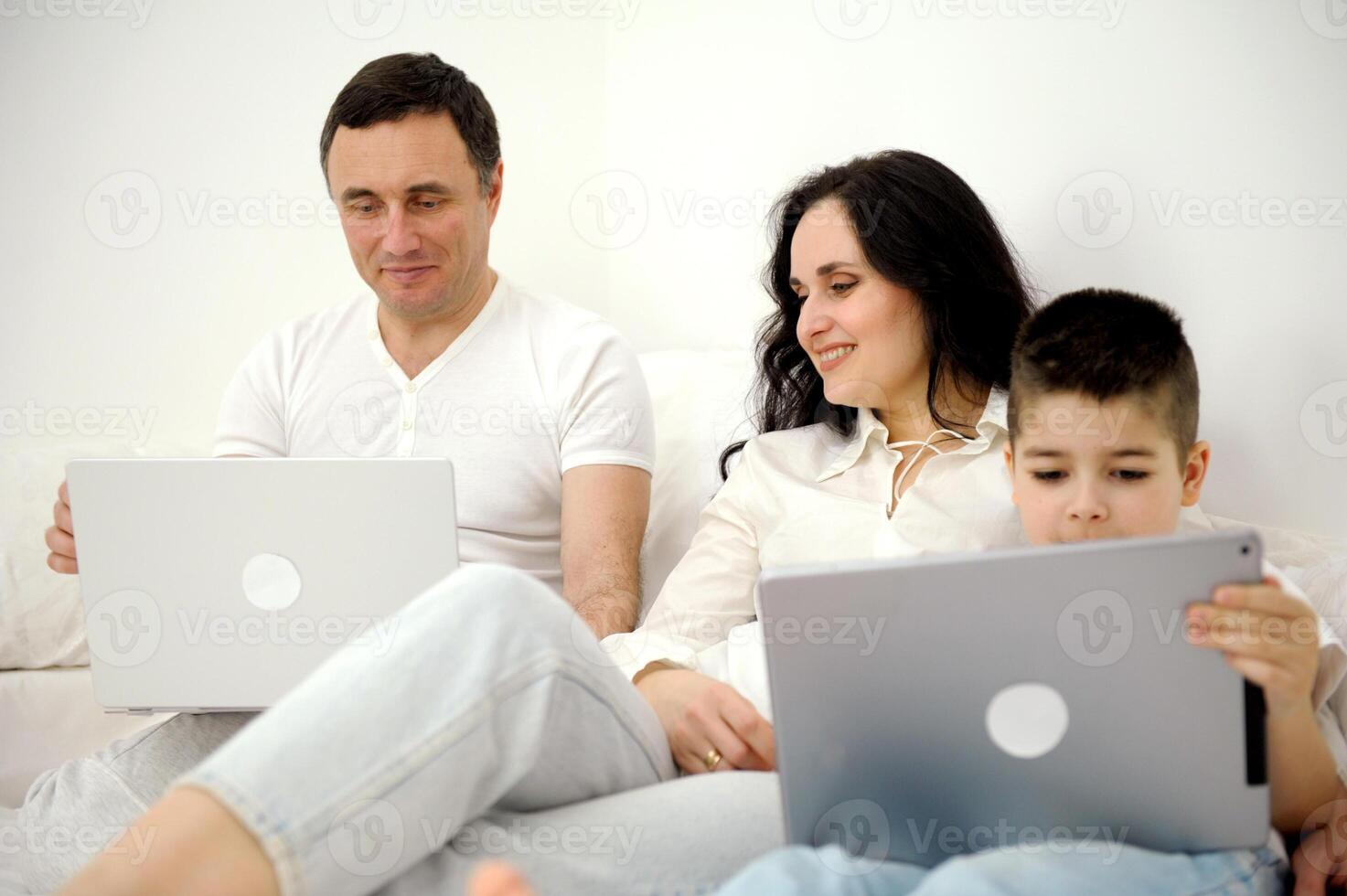 latest technology gadgets mom and son playing on tablet father sitting on white bed working on laptop remote work freelancer vacation family combine work and spend time with family together photo