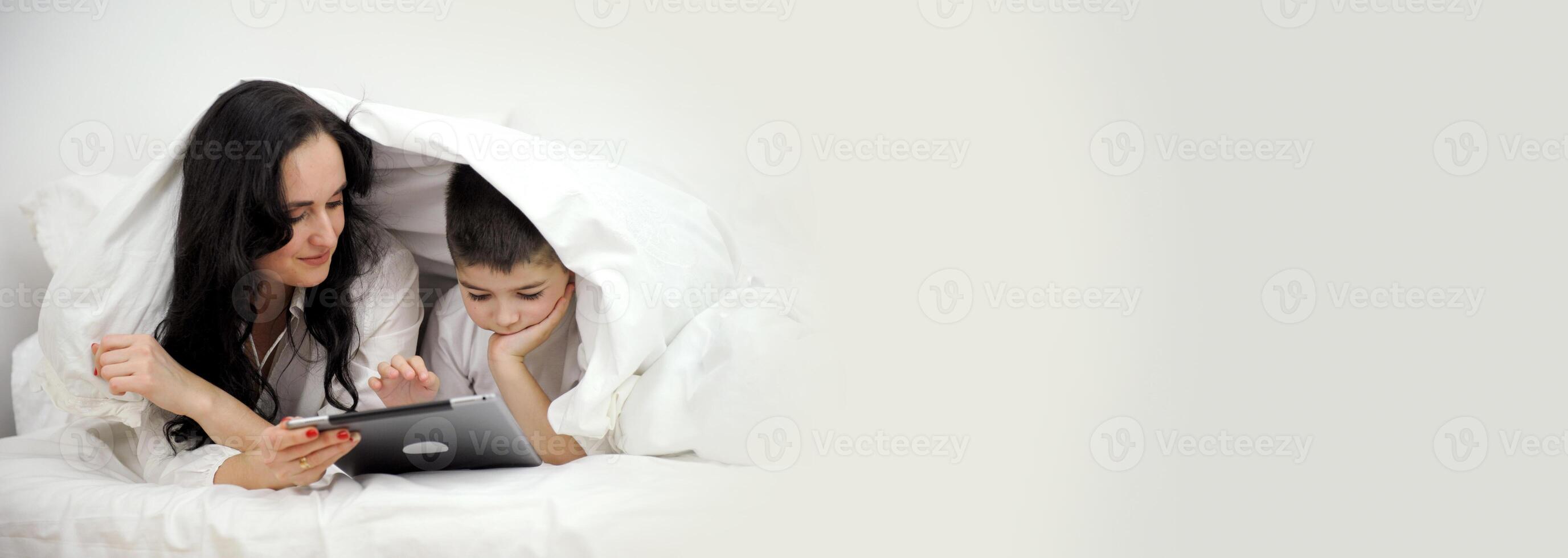 banner family Cosiness mom and son under white duvet playing on a tablet concept heat heating electricity gas apartment payment buying new house construction online study single mom social assistance photo