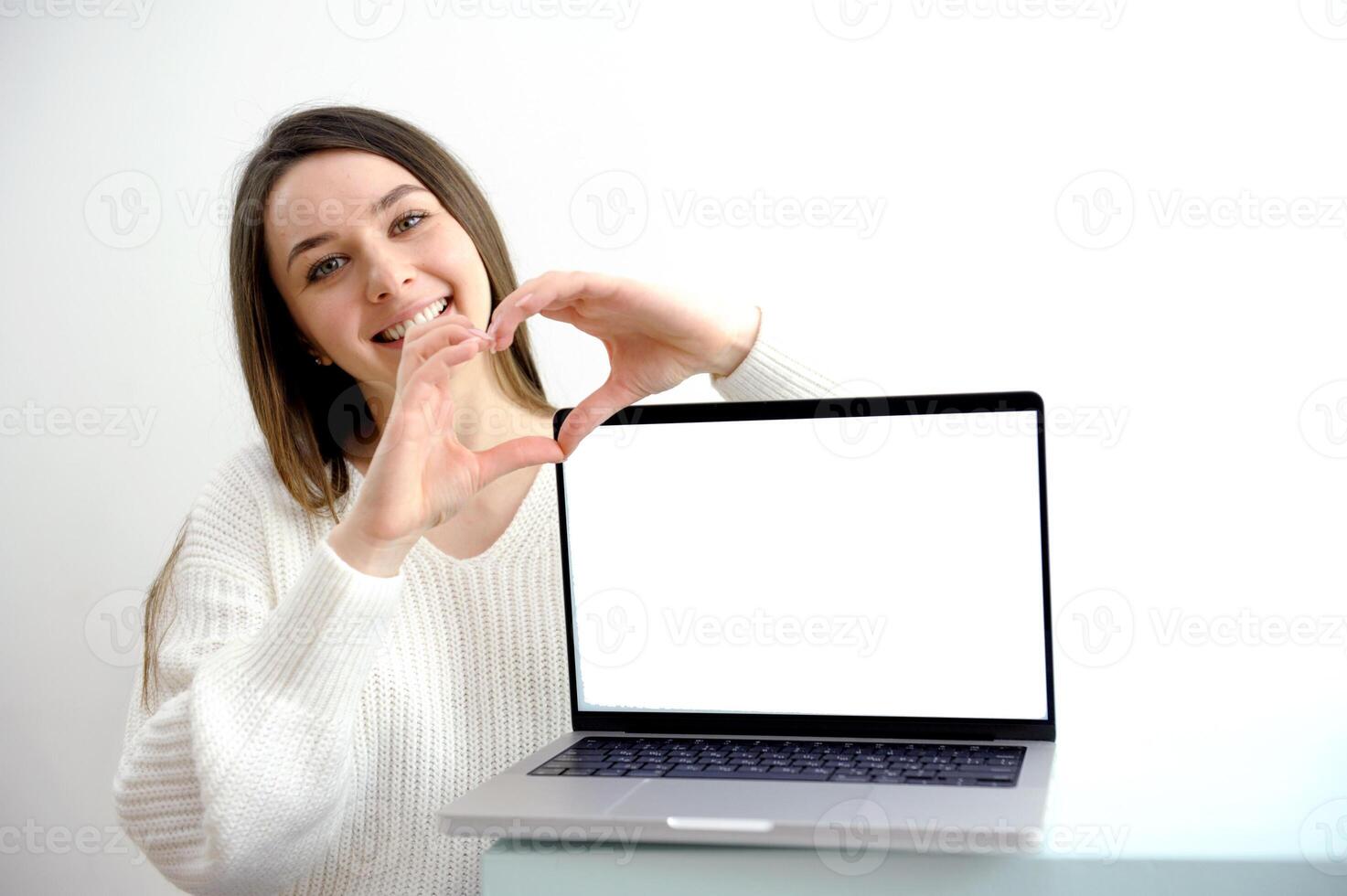 girl on a white background points to a laptop screen a girl in white shows a heart with her hands smiling near a laptop with an empty space for advertising text of any content photo