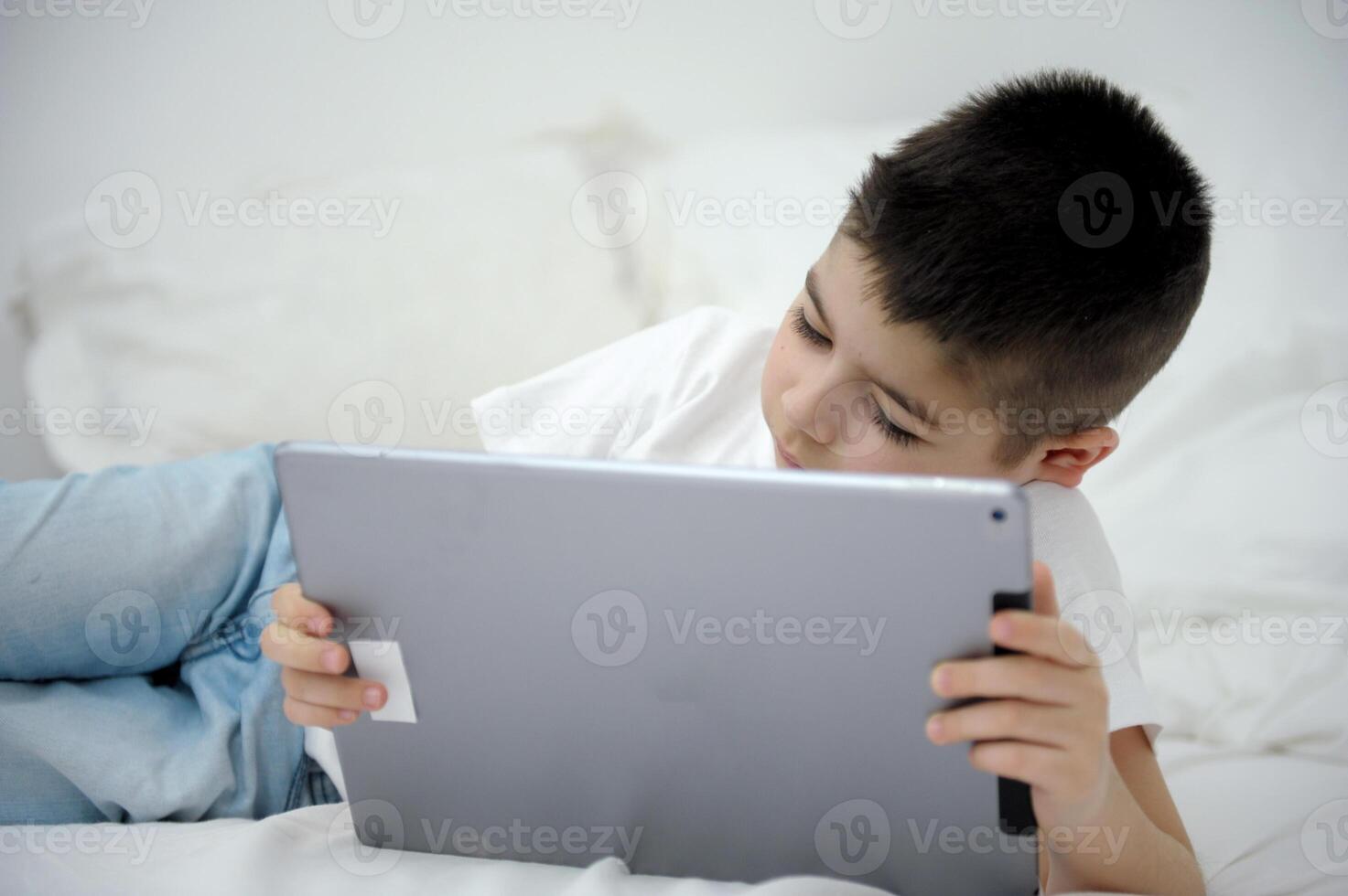 Adorable blond toddler boy laying on the sofa and playing with tablet pc at home, indoors. Child with tablet computer. photo