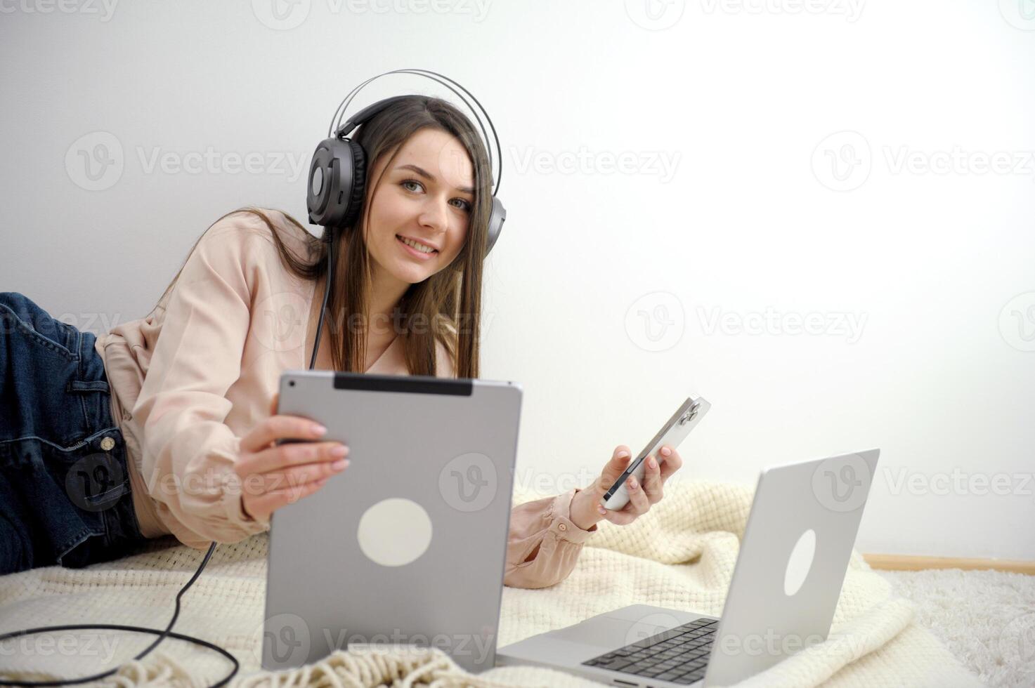 phone tablet laptop a lot of different technologies girl listens to music headphones plays phone works on computer on floor bed rest at home Work online on Internet social networks freelancer photo