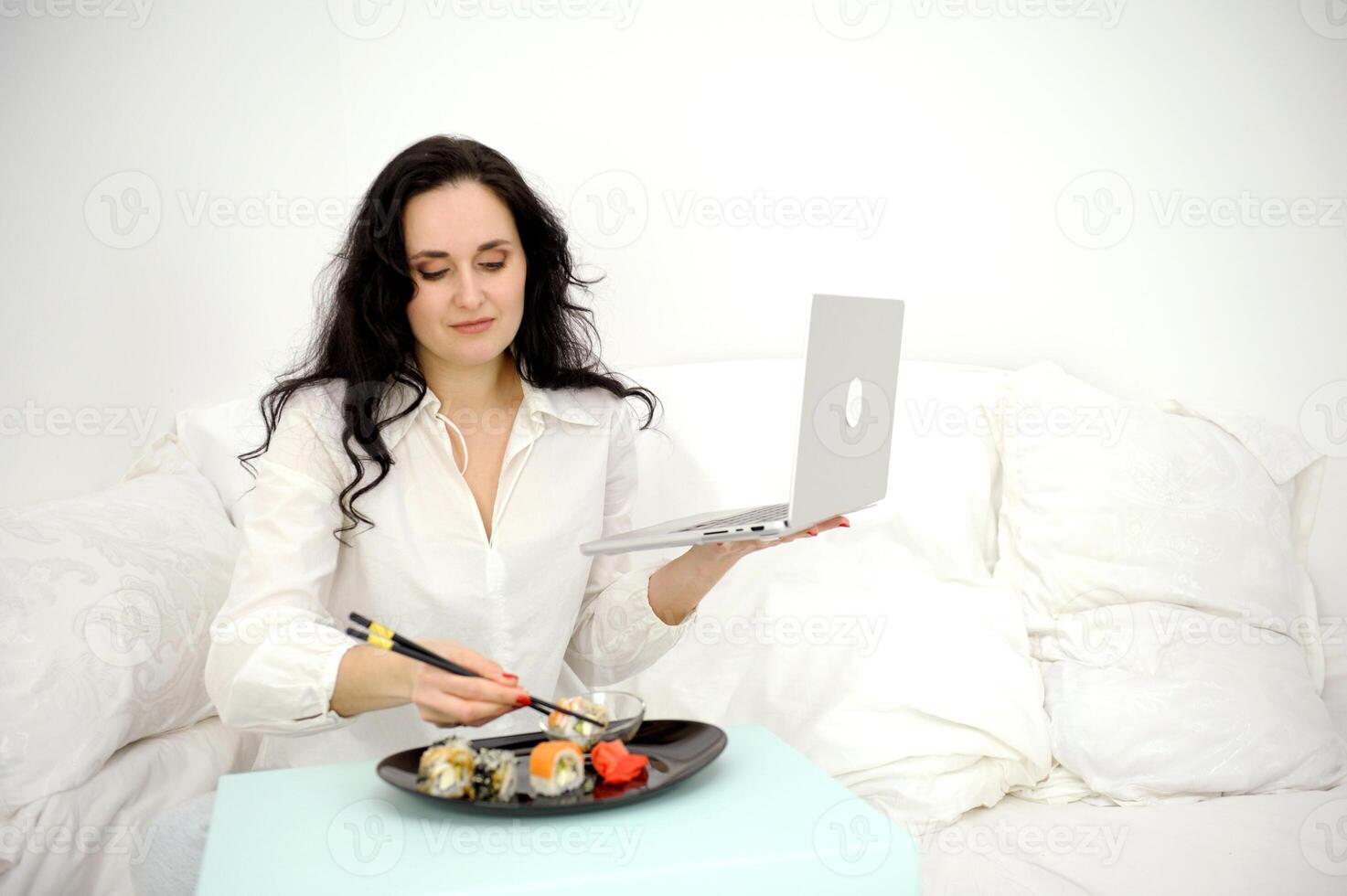 Pleasant caucasian lady sit on bed eating sushi while watching movie at home. Adorable short haired female in domestic wear have rest at weekends, holidays. leisure, relaxation, lifestyle photo