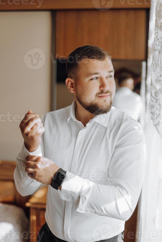 A man in a white shirt is getting dressed and adjusting his watch photo