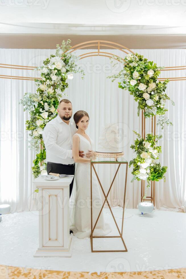 A bride and groom stand in front of a cake on a table photo