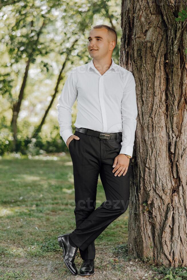 A man in a white shirt and black pants stands next to a tree photo