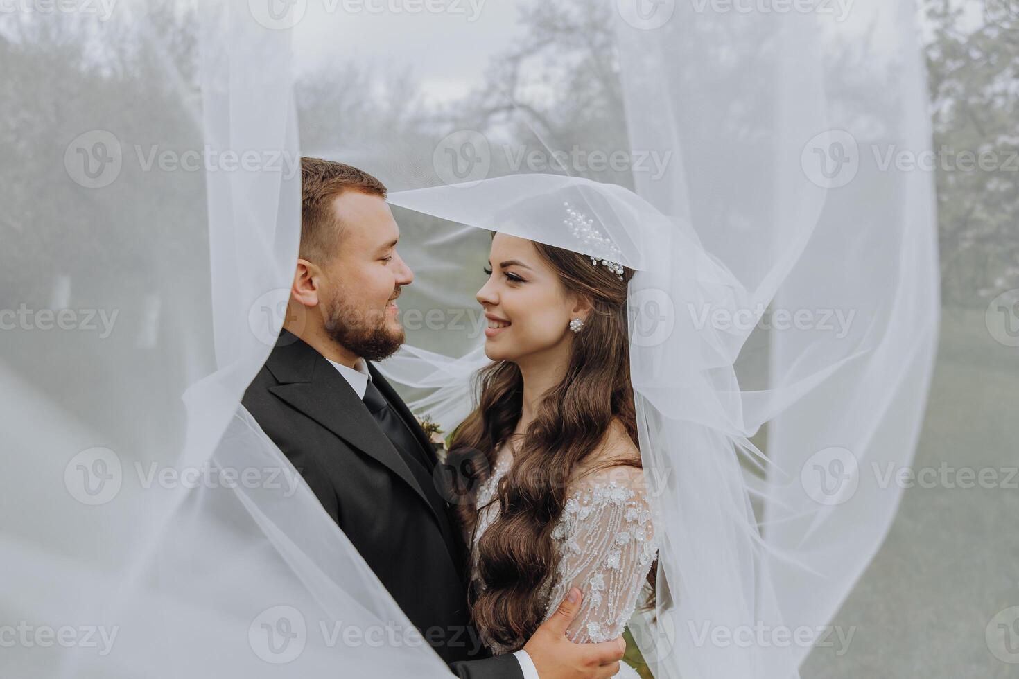 a handsome and stylish groom embraces his bride in a white dress under her veil photo