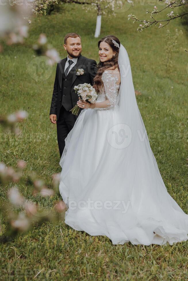 the groom and his bride are walking on the green grass in the spring garden. The bride is in a chic white dress, the groom is in a black suit photo