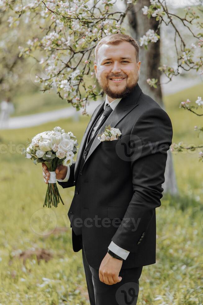 The groom, dressed in a black suit, stands in nature with a bouquet of flowers in his hand. An elegant man in a classic black suit. Portrait of a man photo