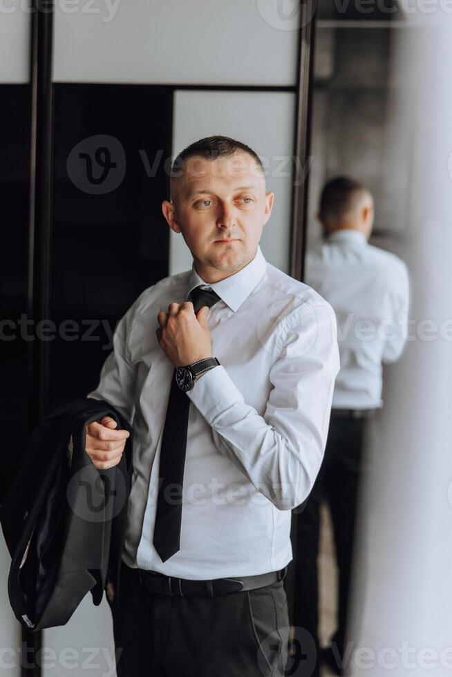A man in a suit and tie is standing in front of a mirror, adjusting his tie photo