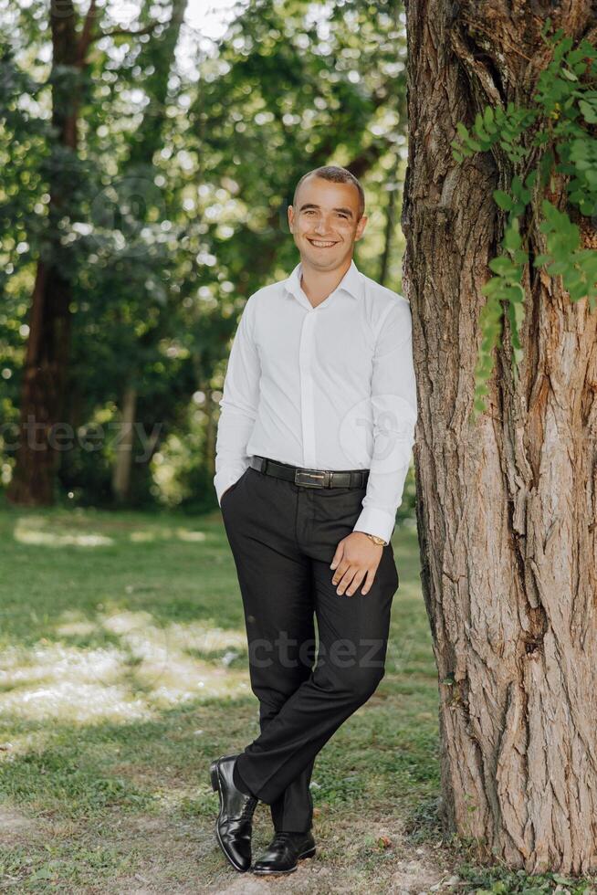 A man in a white shirt and black pants stands next to a tree photo