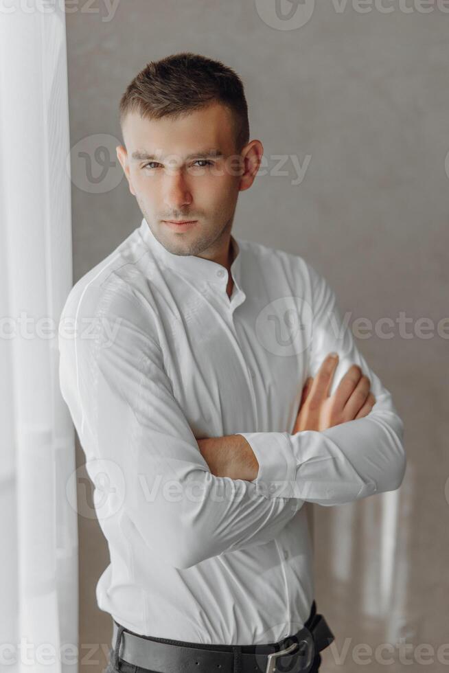 A man in a white shirt is standing in front of a window, with his arms crossed photo