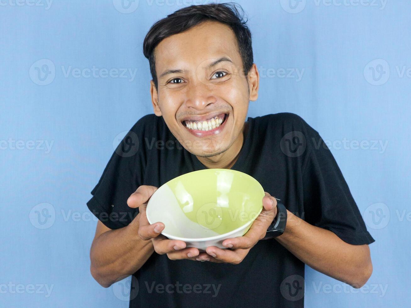 A young Asian man in a black t-shirt stands and holds an empty bowl showing the dish. photo