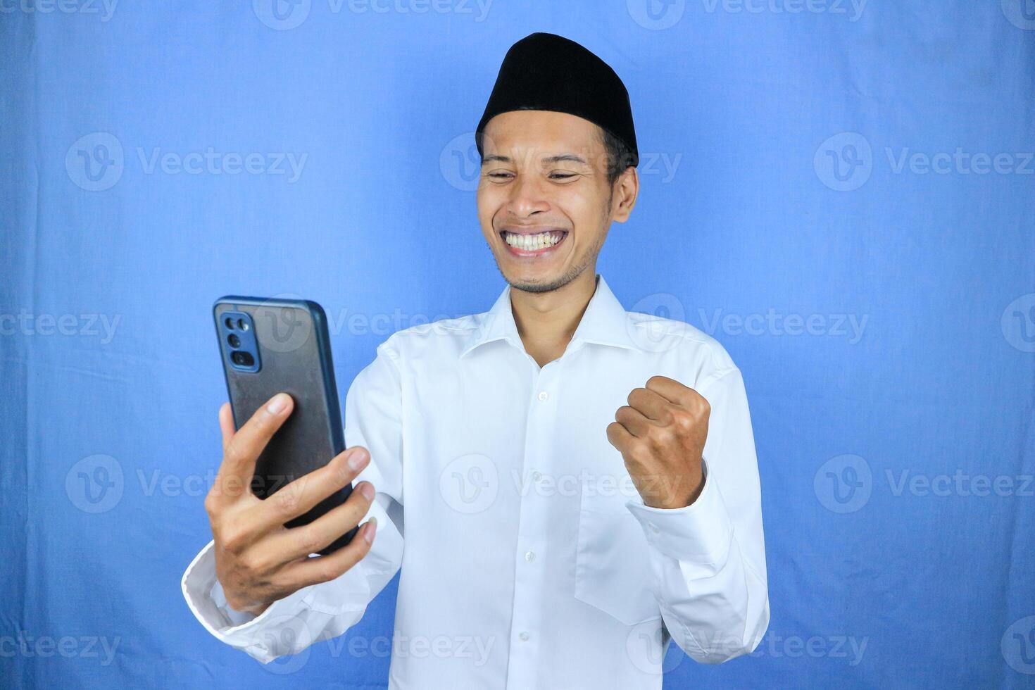 Muslim asian man showing excited expression with fist clenched while holding mobile phone photo