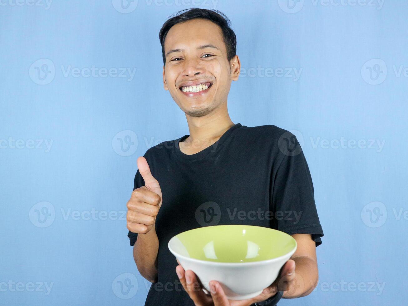 A young Asian man in a black t-shirt stands and holds an empty bowl showing the dish. photo