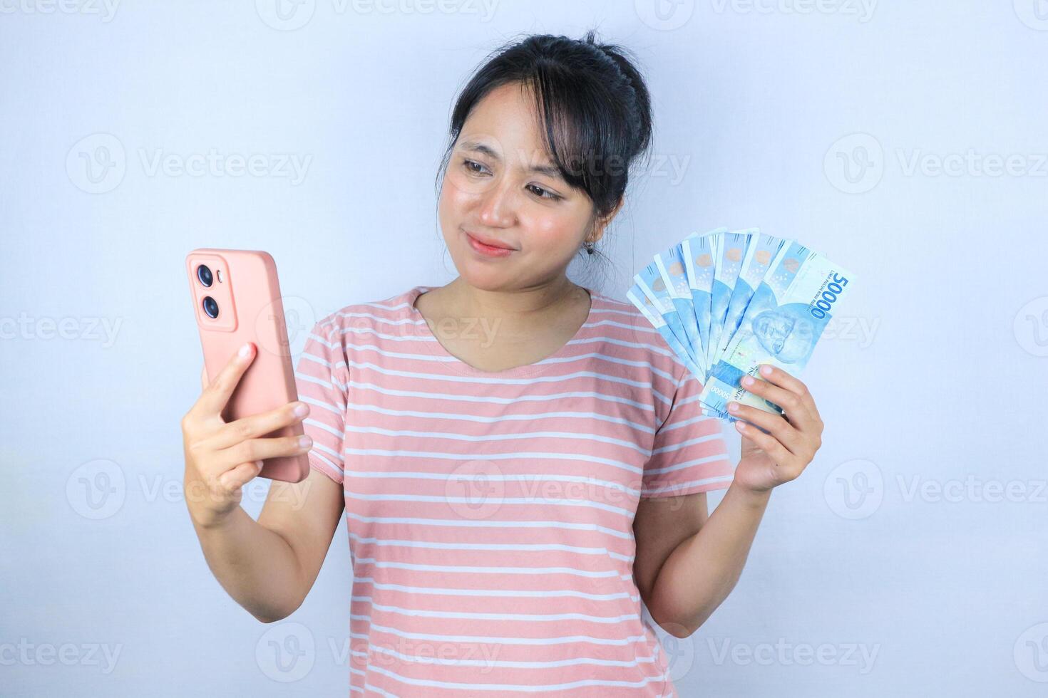 smiling expression of asian woman holding cash rupiah while looking at phone screen. photo