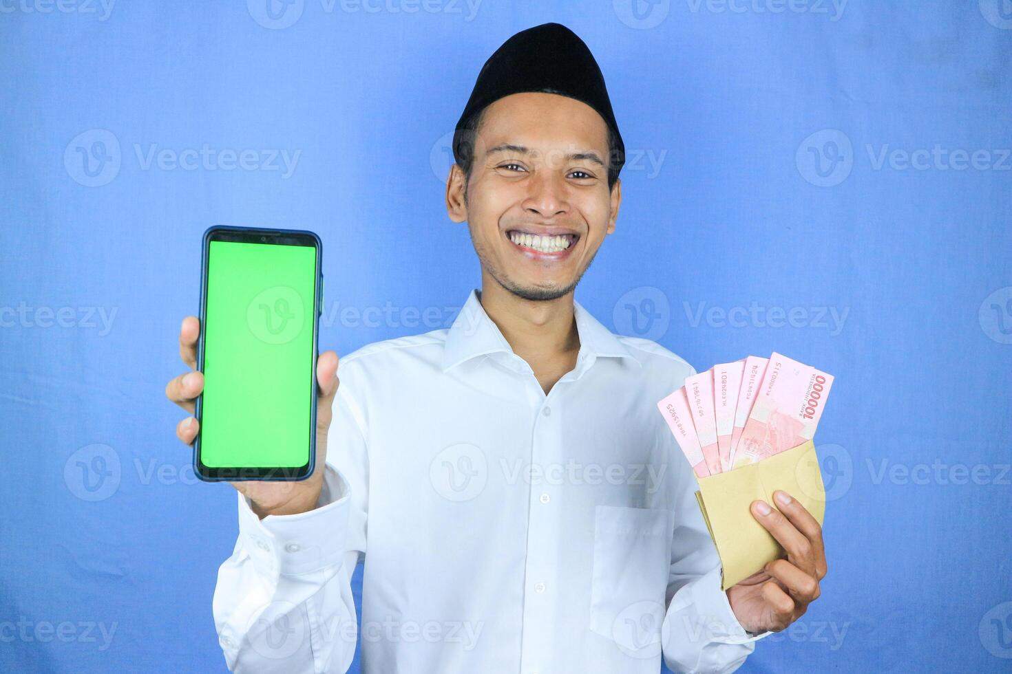 Happy expression of Asian Muslim man holding money rupiah banknotes while showing empty phone screen photo