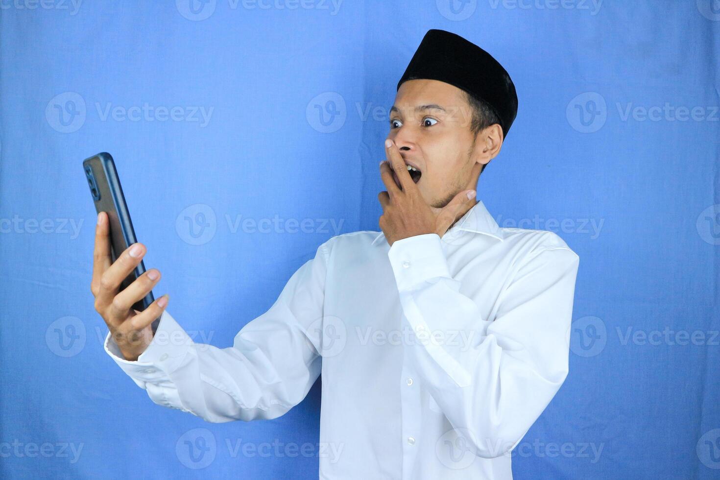 funny facial expression of asian muslim man wearing cap shocked looking at phone screen photo