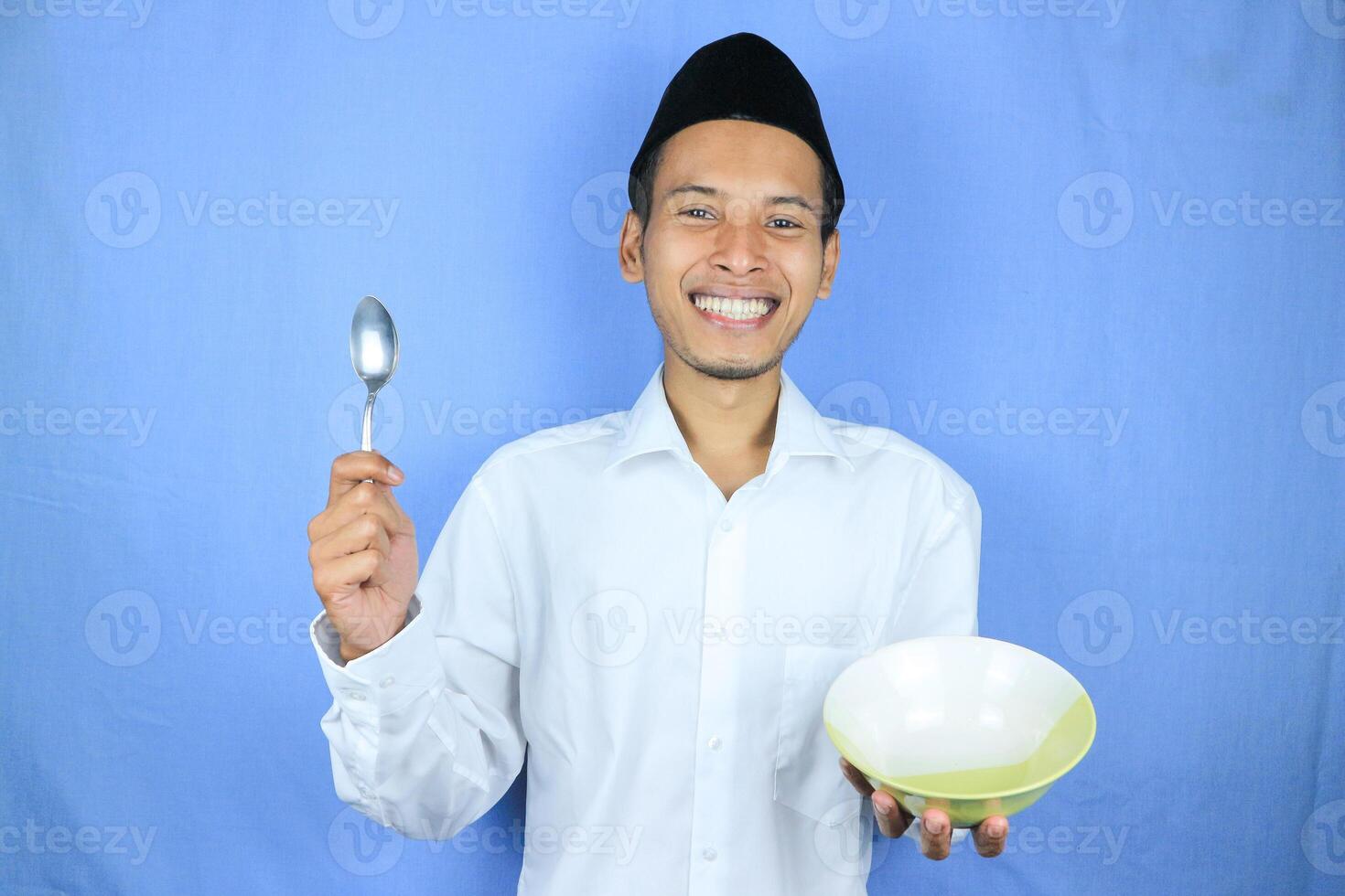Muslim Asian man wearing cap stand and hold an empty bowl and spoon showing the dish photo