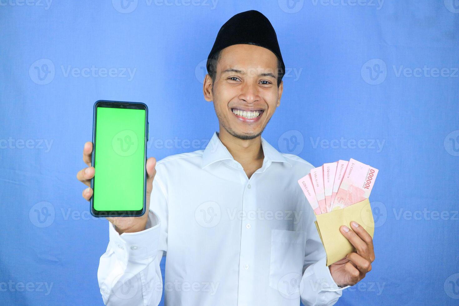 Happy expression of Asian Muslim man holding money rupiah banknotes while showing empty phone screen photo
