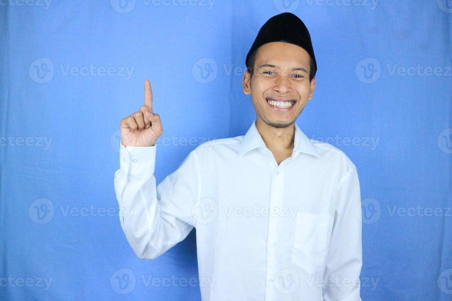 Smiling expression of muslim asian man, pointing finger an empty space. Advertising concept photo
