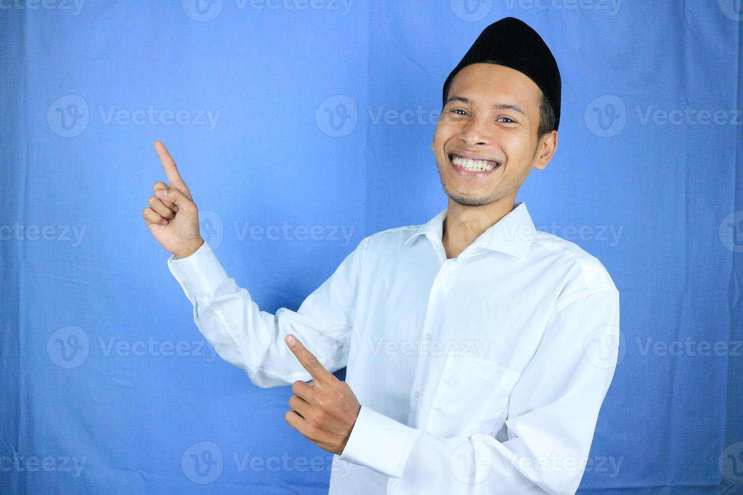 Smiling expression of muslim asian man, pointing finger an empty space. Advertising concept photo