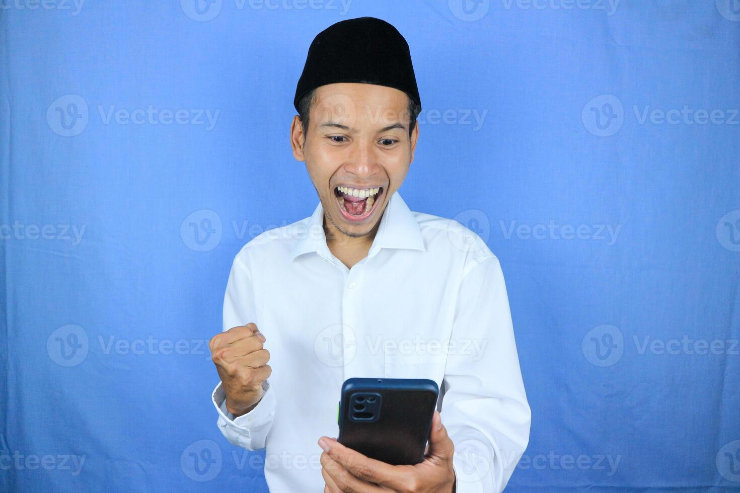 Muslim asian man showing excited expression with fist clenched while holding mobile phone photo