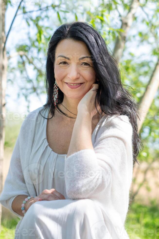 Smiling woman, middle aged, with long dark hair, wearing a white blouse, enjoying a sunny nature setting. Exudes a serene vibe of self care and well being. photo