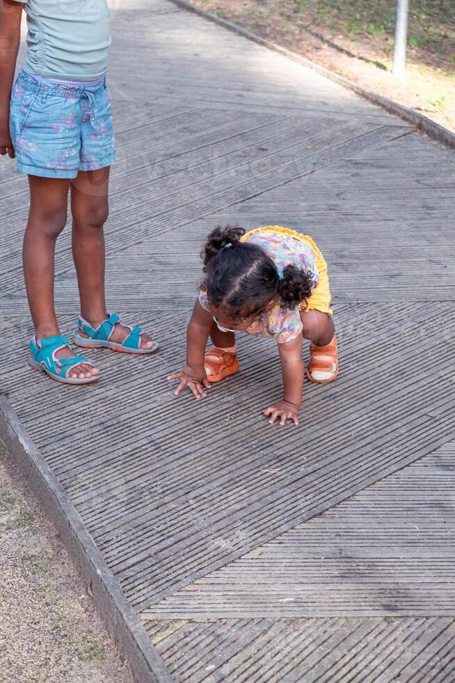 A black toddler crouches on a wooden boardwalk, peering down, beside a standing child in blue shorts. The scene is innocent, ideal for family or park themes. photo