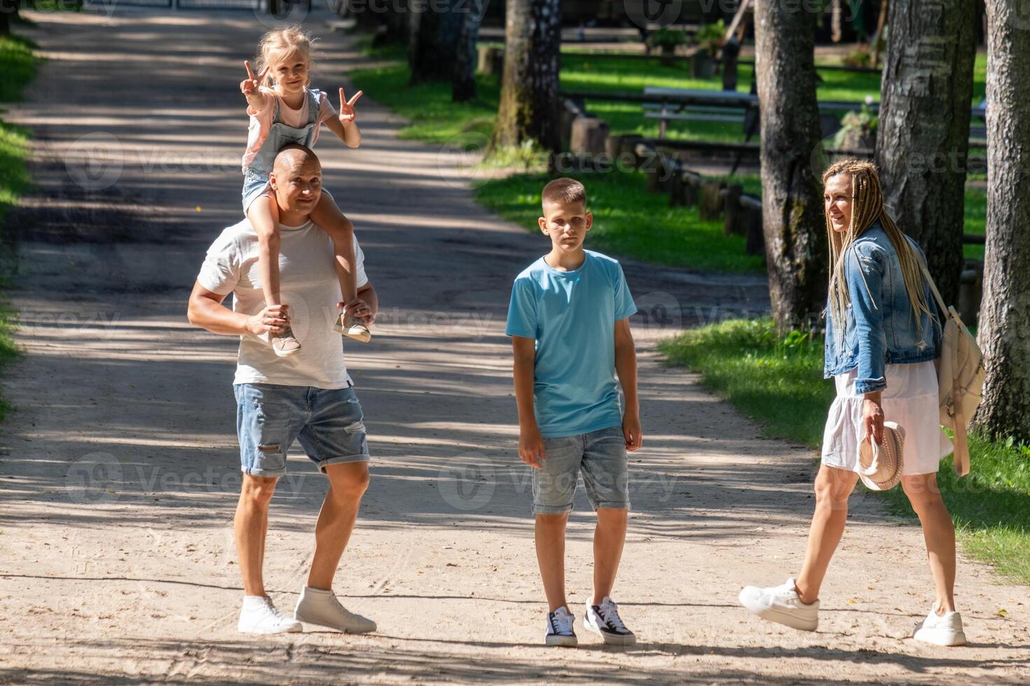 A family enjoys a walk in a sun-dappled park a father with a child on his shoulders, another child, and their mother, embodying the joy of family bonding, National Siblings Day. photo