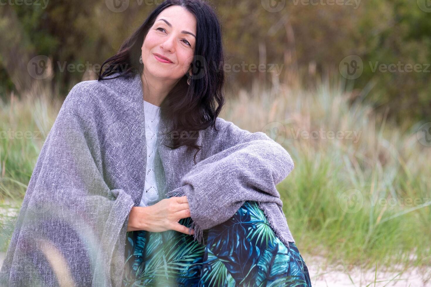 A serene middle aged woman wrapped in a cozy gray shawl sits outdoors, exuding tranquility amidst a natural backdrop, suggesting contemplation and life's autumnal wisdom. photo