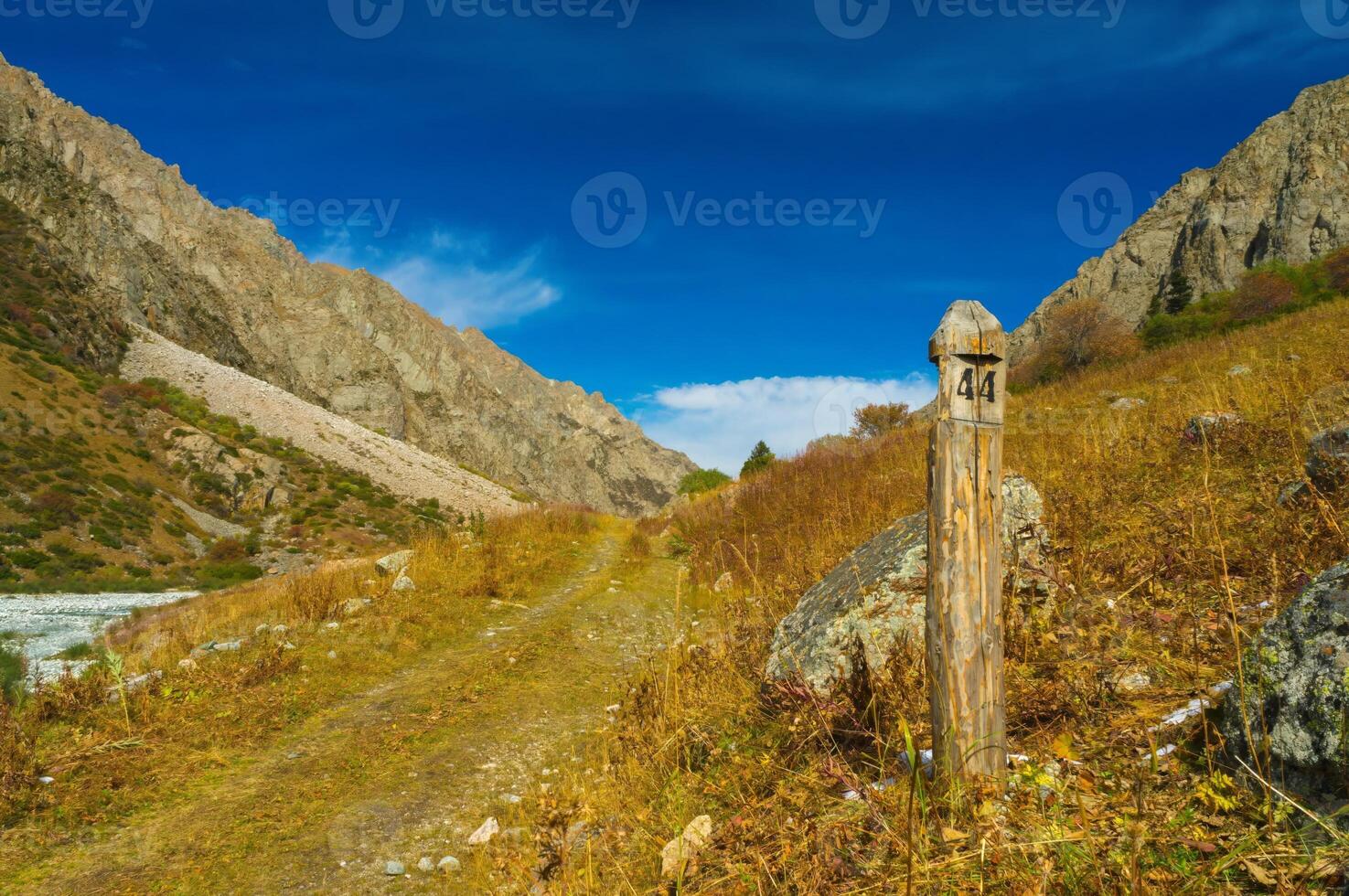 A rustic wooden milepost stands by a meandering path through a golden valley framed by imposing grey mountains, under a vast blue sky with scattered clouds, evoking tranquility and remoteness. photo