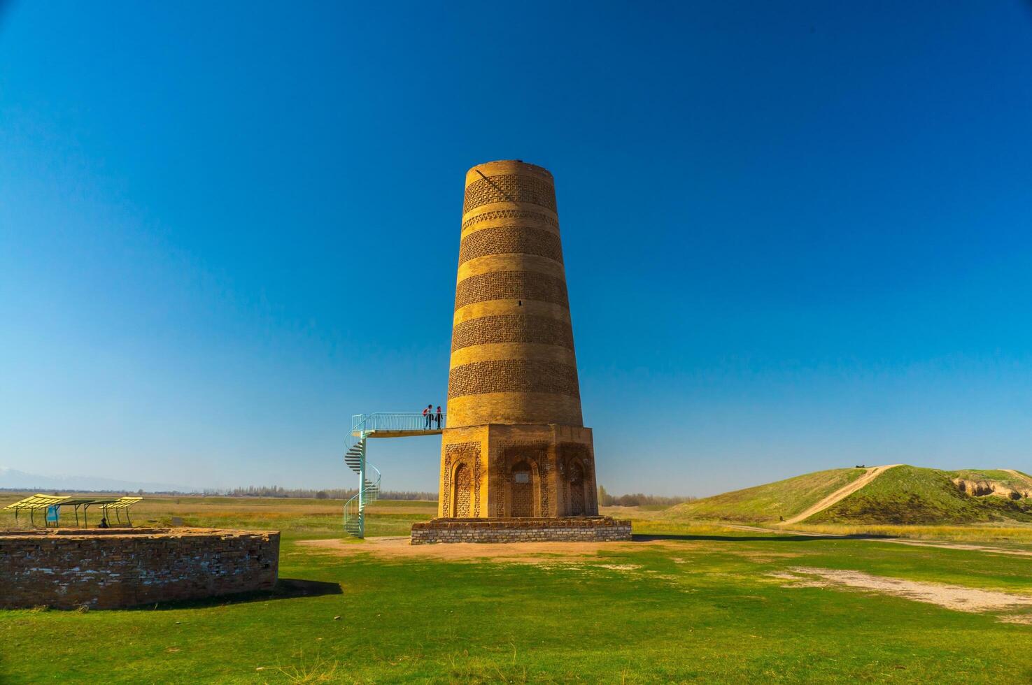 The Burana Tower in Kyrgyzstan, an 11th-12th century minaret, featuring ascending visitors on its external staircase, towers over green fields under a vast blue sky. photo