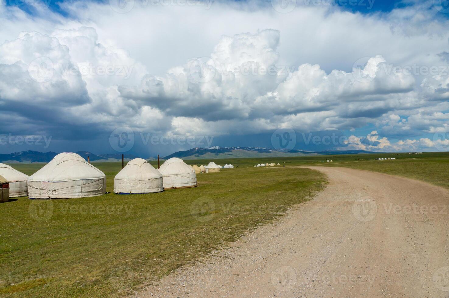 Row of traditional Kyrgyz yurts dot the open grasslands of Son-Kul, with distant mountains framing the horizon, under a vast sky marked by streaks of white clouds, reflecting a nomadic heritage. photo