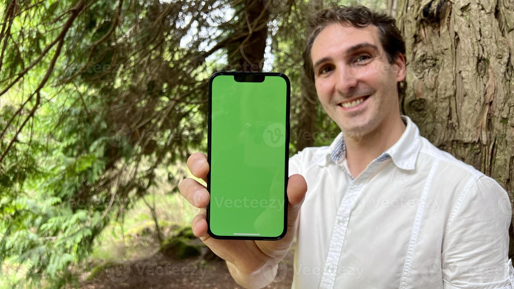 very handsome man french italy not holding a phone with a green tap standing in the woods in a white shirt and smiling showing a finger on the screen can use for advertising photo