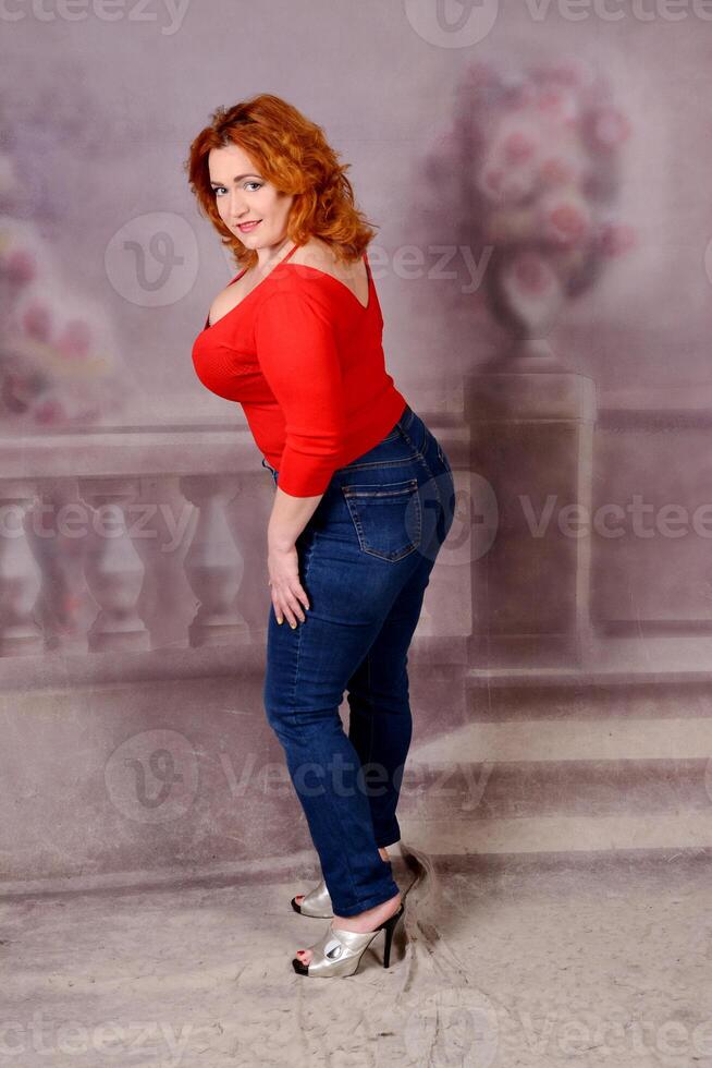 Redhead caucasian girl in red pattern blouse and jeans smiles staying near grey background photo