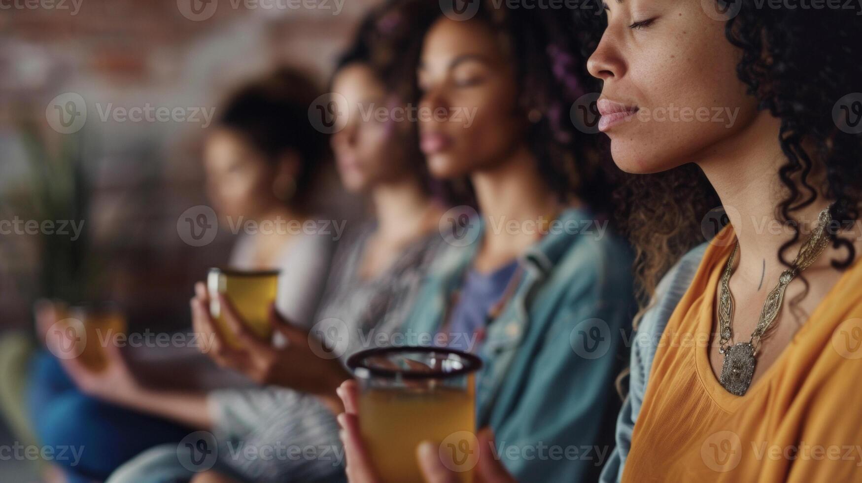 A group of women sit in a circle their eyes closed as they meditate and chant together. Each woman is holding a different herbal tea drank before or after the yoga practic photo