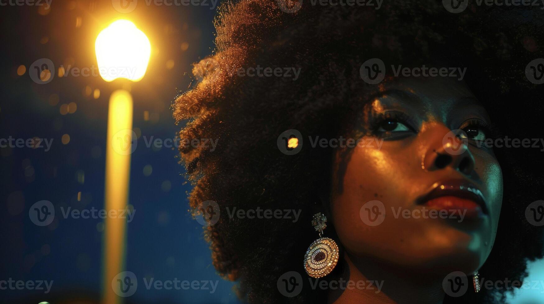 Illuminated by a single streetlight a black woman embraces her natural hair and features adorned with sparkling jewels and deep mystical makeup. As she stares into the distance it photo