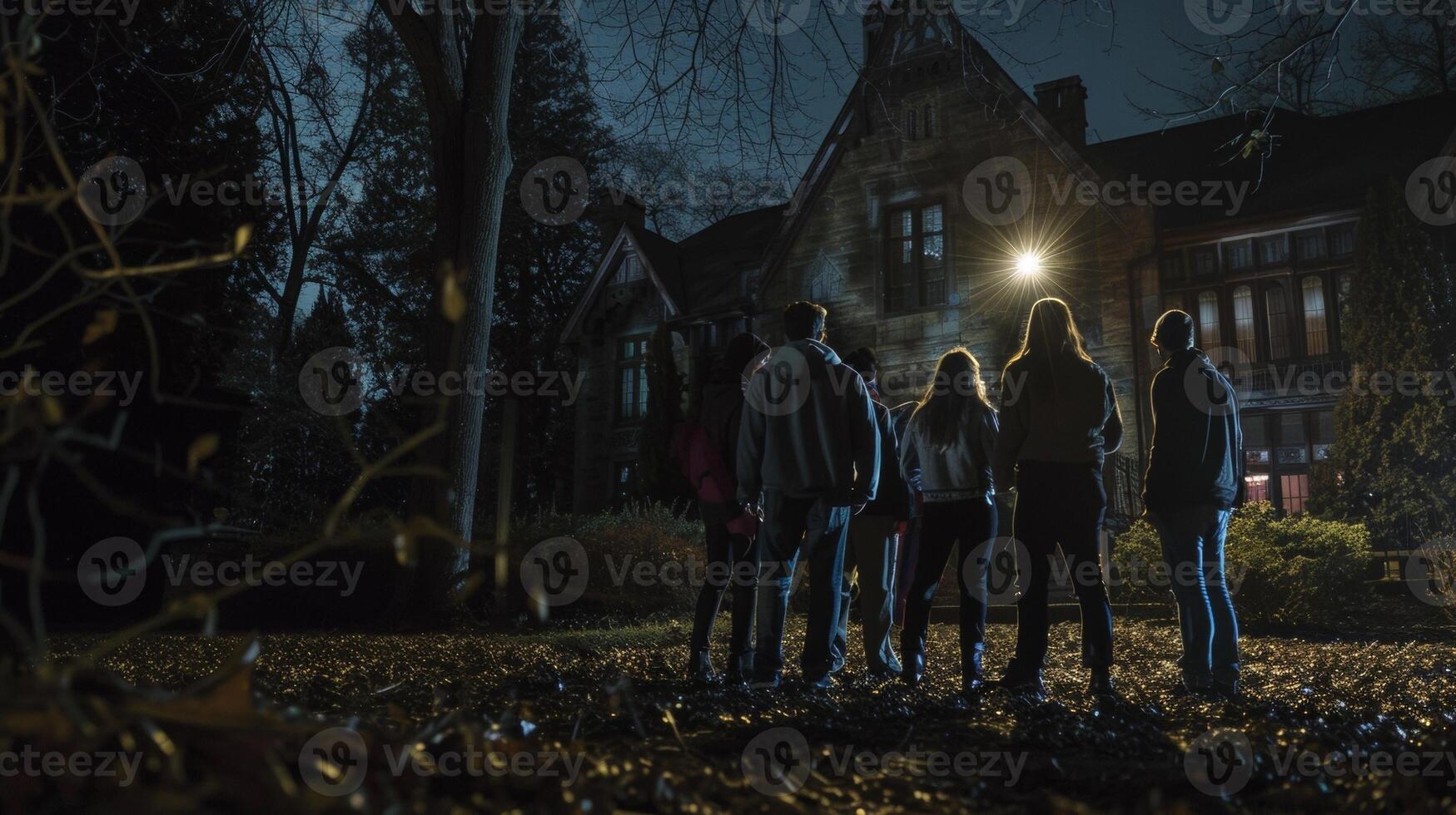 A group of adults stand in a huddle backs facing the camera as they survey the ominous mansion ahead. One man can be seen holding . photo